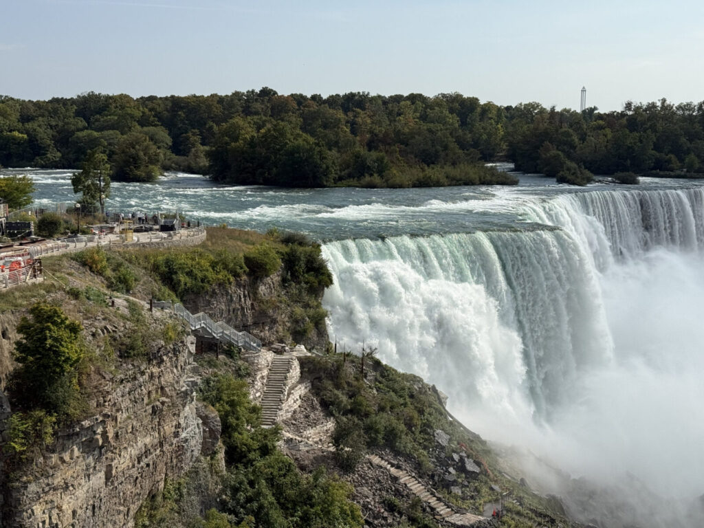 View of Niagara Falls with water cascading over the edge, surrounded by greenery and a rocky cliffside staircase.