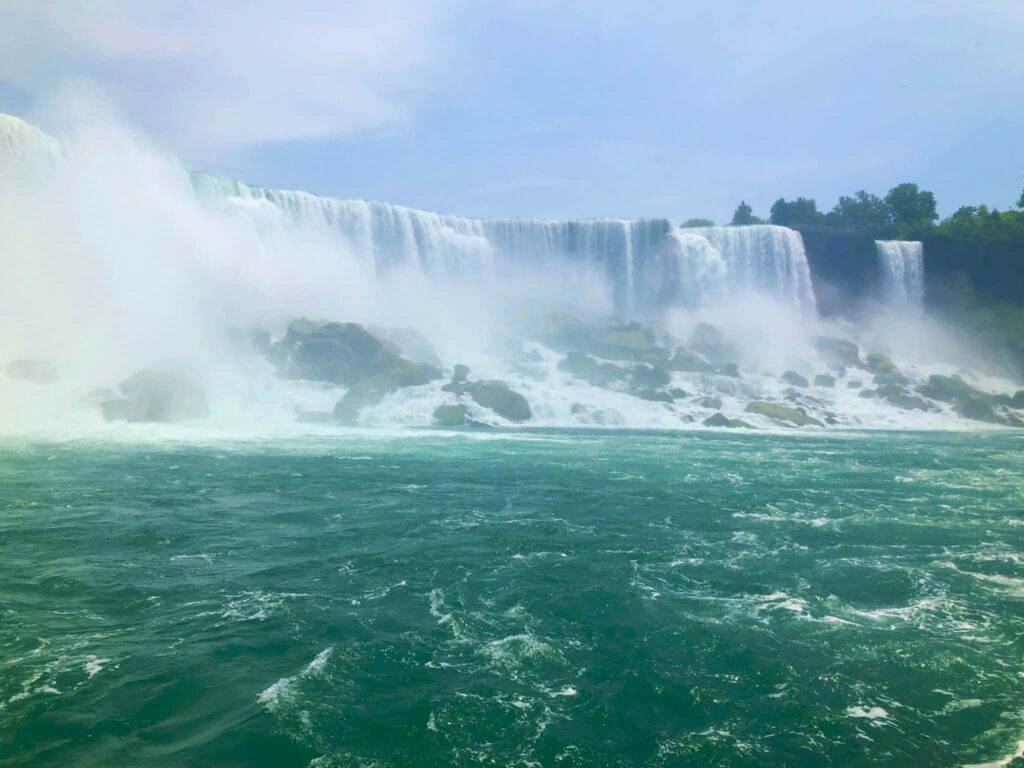 Wide waterfall cascades over rocky cliffs into a turquoise river, with mist rising and trees in the background.