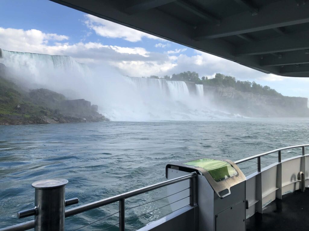 View of a waterfall and mist from the deck of a boat, with a railing and part of the boat visible in the foreground.