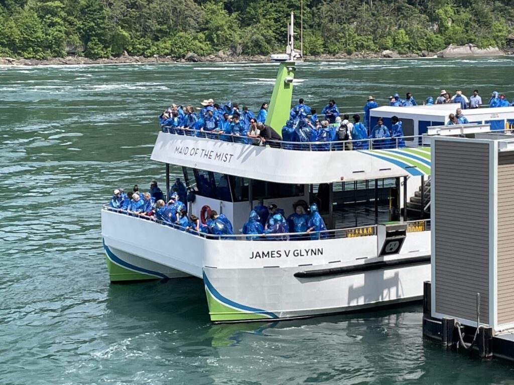 A crowded Maid of the Mist tour boat named James V Glynn carries passengers in blue rain ponchos near a dock.